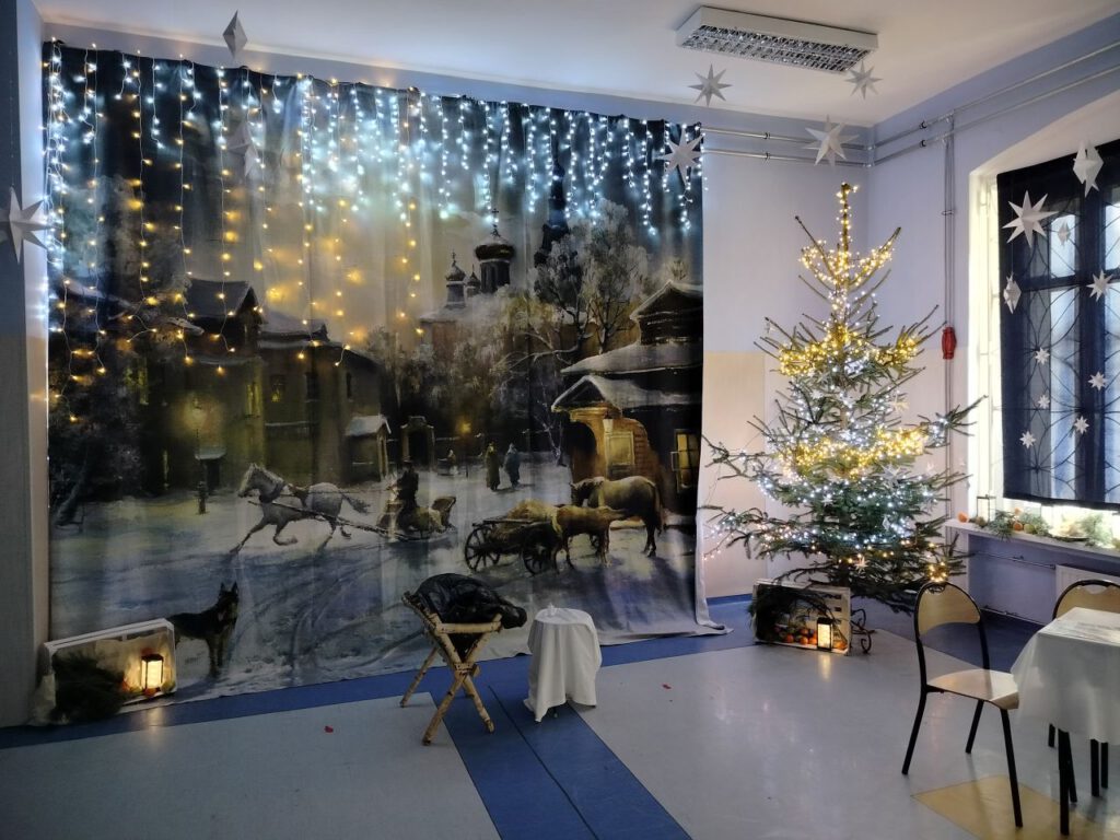 Dekoracja sali na Jasełka, na tle dużego obrazu przedstawiającego zimowy krajobraz stoi żłóbek.