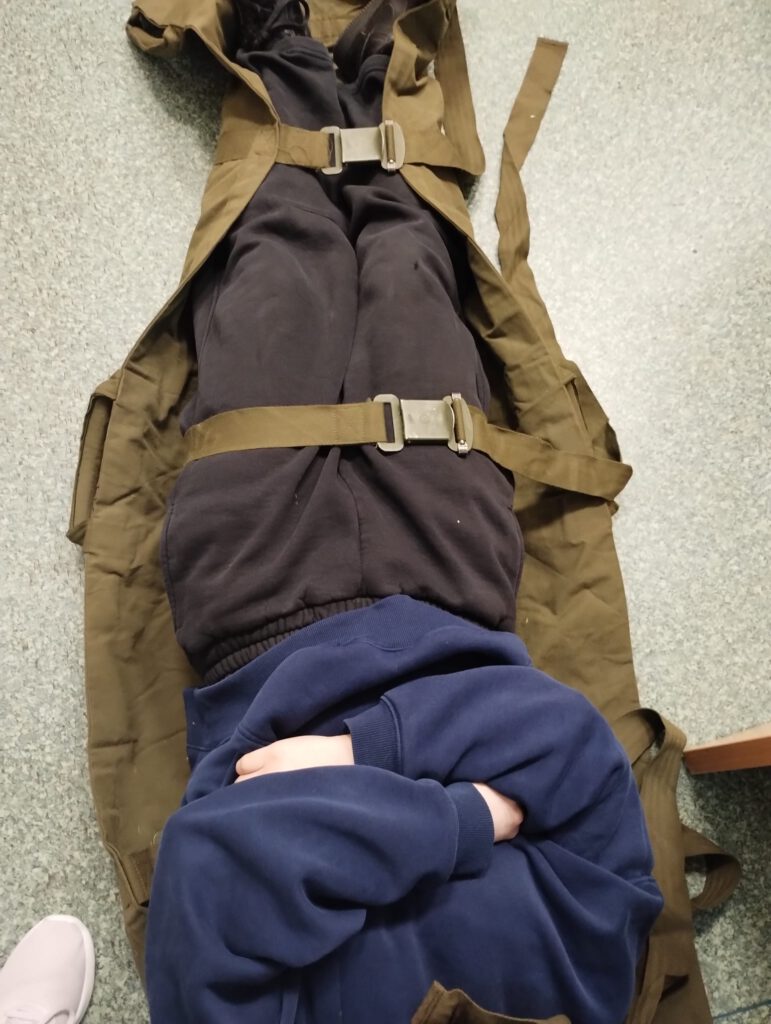 Na zdjęciu przedstawiono sposób zabezpieczenia poszkodowanego w noszach wojskowych.